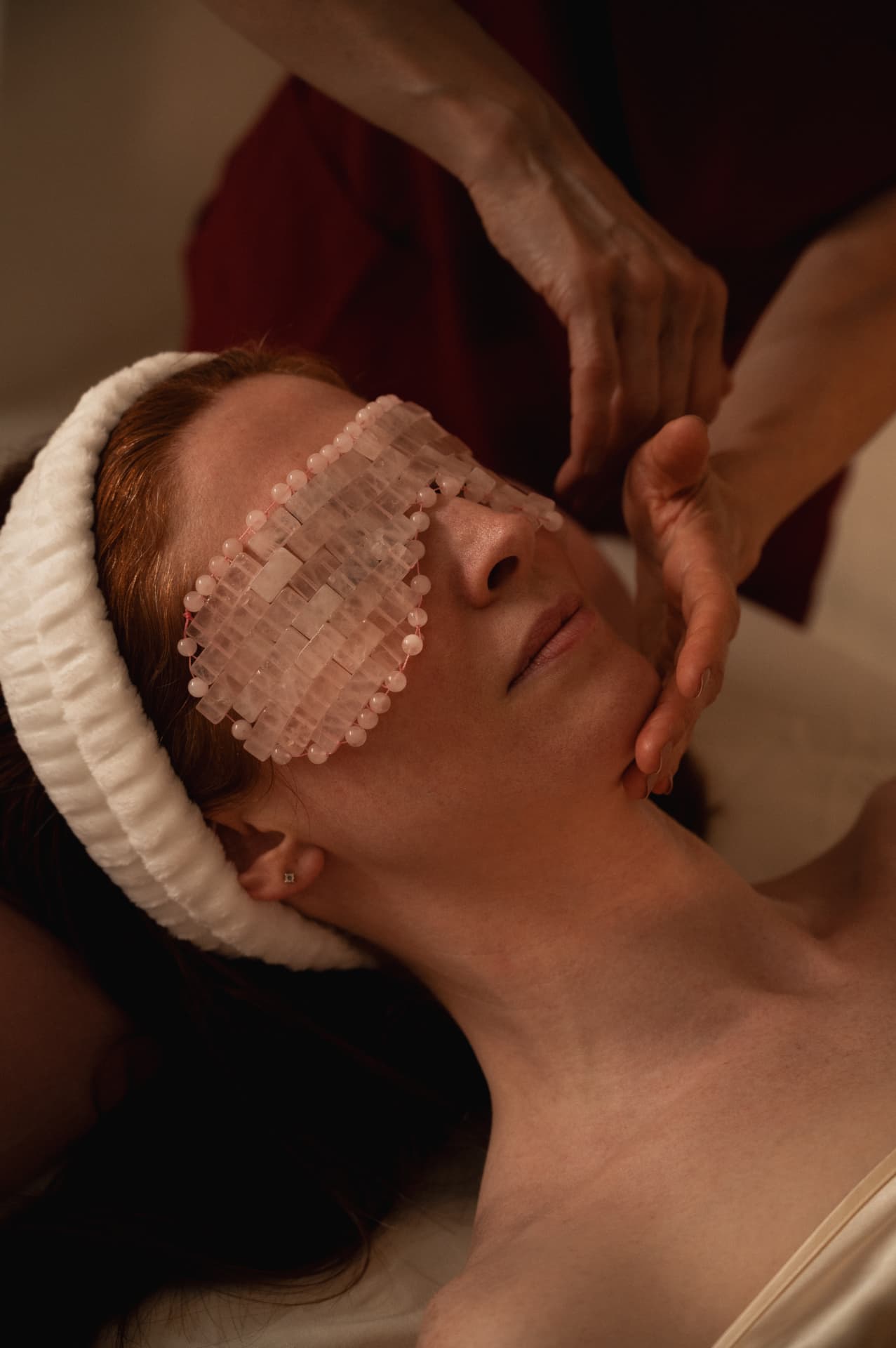 A rose quartz eye mask resting over a woman eyes while a practitioner works slowly along her jaw and cheek