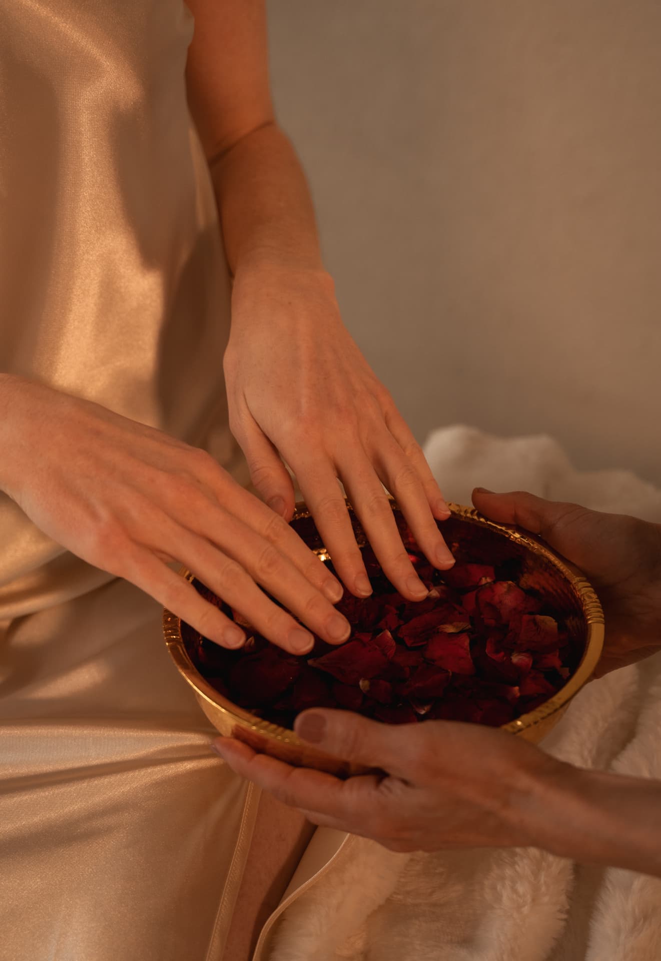 A woman in champagne silk lowering both hands into a gold bowl of dark red rose petals