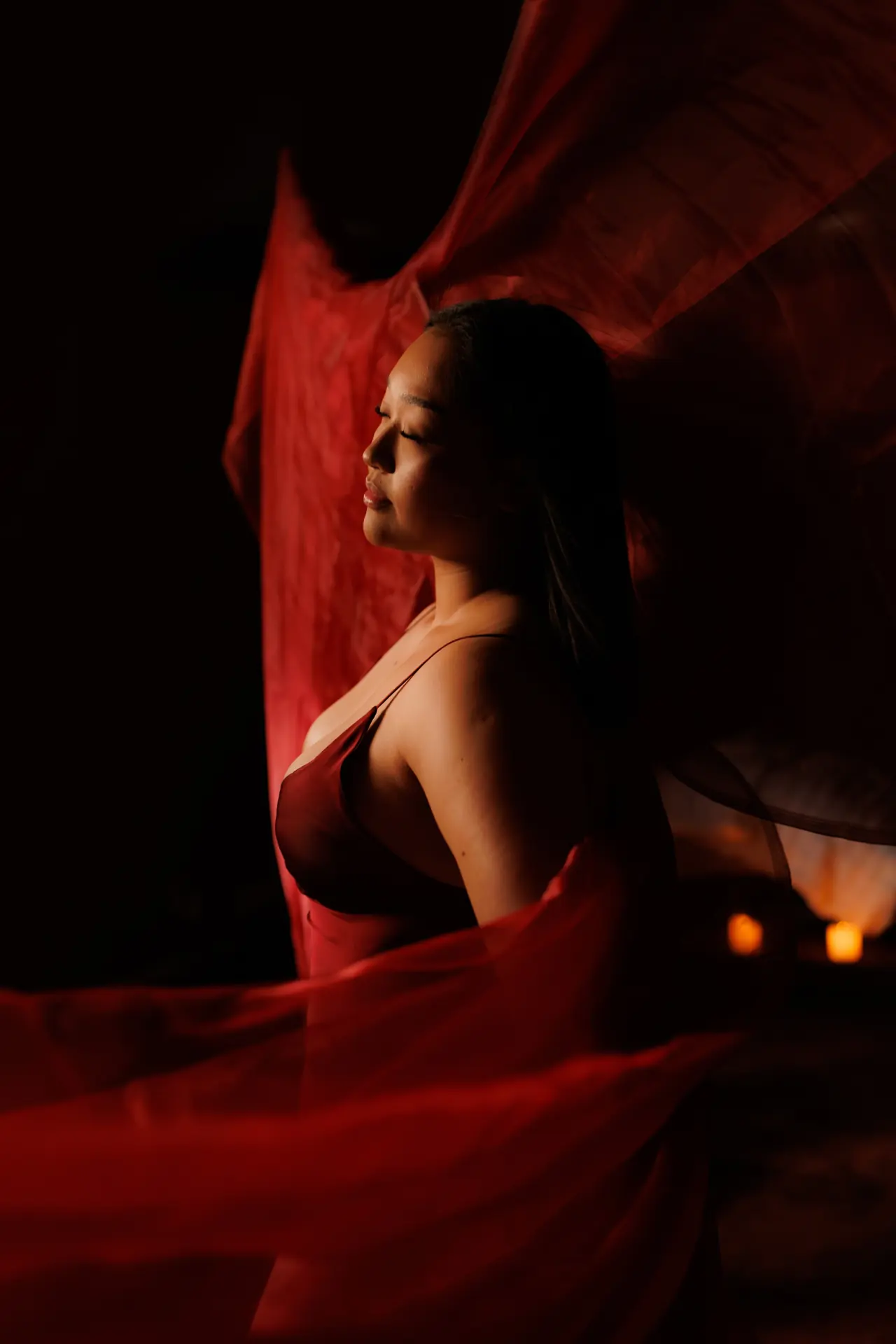 A woman with her eyes closed in a dim room, deep red fabric drifting behind her and candles burning low nearby
